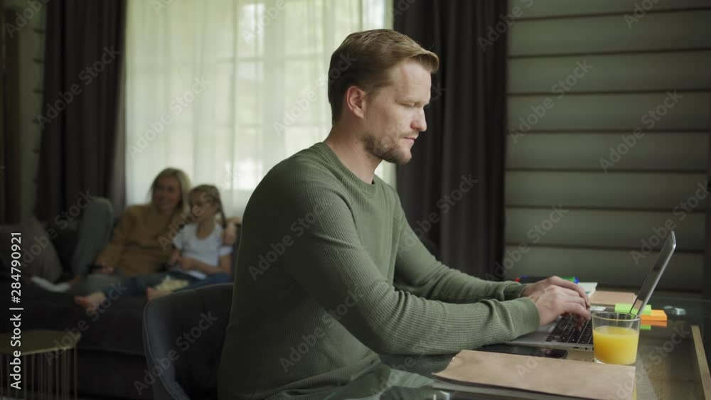 Tracking right shot of entrepreneur man working on laptop computer sitting at desk at home, his wife and little daughter watching tv in background