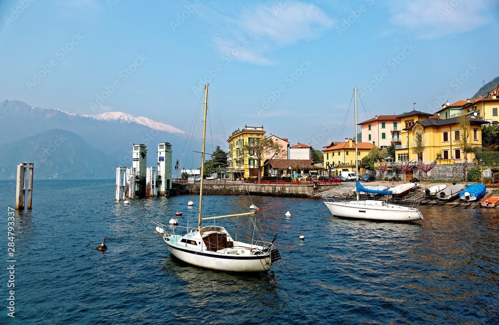 Lakeside scenery of Varenna, a beautiful village by Lake Como in Lombardy, Italy with view of sailboats parking at the pier, colorful houses by lakeshore & snow capped mountains in distant background