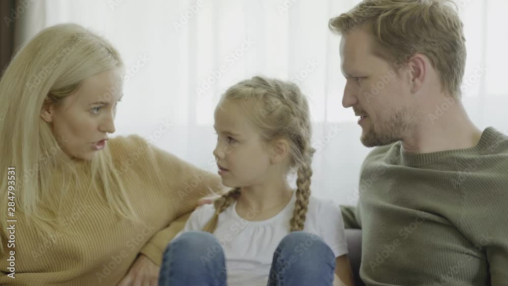 Beautiful family of mother, father and little daughter sitting on sofa at home and talking, medium shot Beautiful family of mother, father and little daughter sitting on sofa at home and talking, medium shot