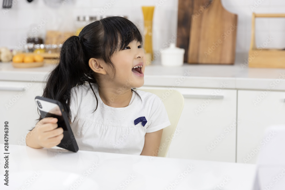 Happy Asian Little girl enjoy smart phone while waiting for the mother preparing breakfast in the kitchen at home. Technology and everyday life Happy Asian Little girl enjoy smart phone while waiting for the mother preparing breakfast in the kitchen at home. Technology and everyday life