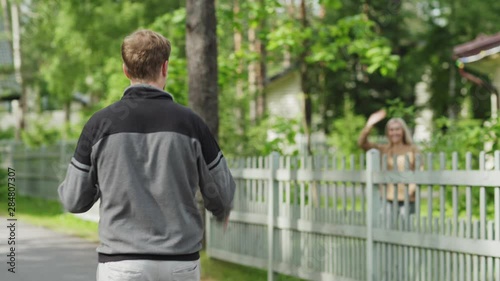Man walking home in countryside and texting on cell phone. Male meeting smiling female neighbor, greeting her and talking cheerfully over fence