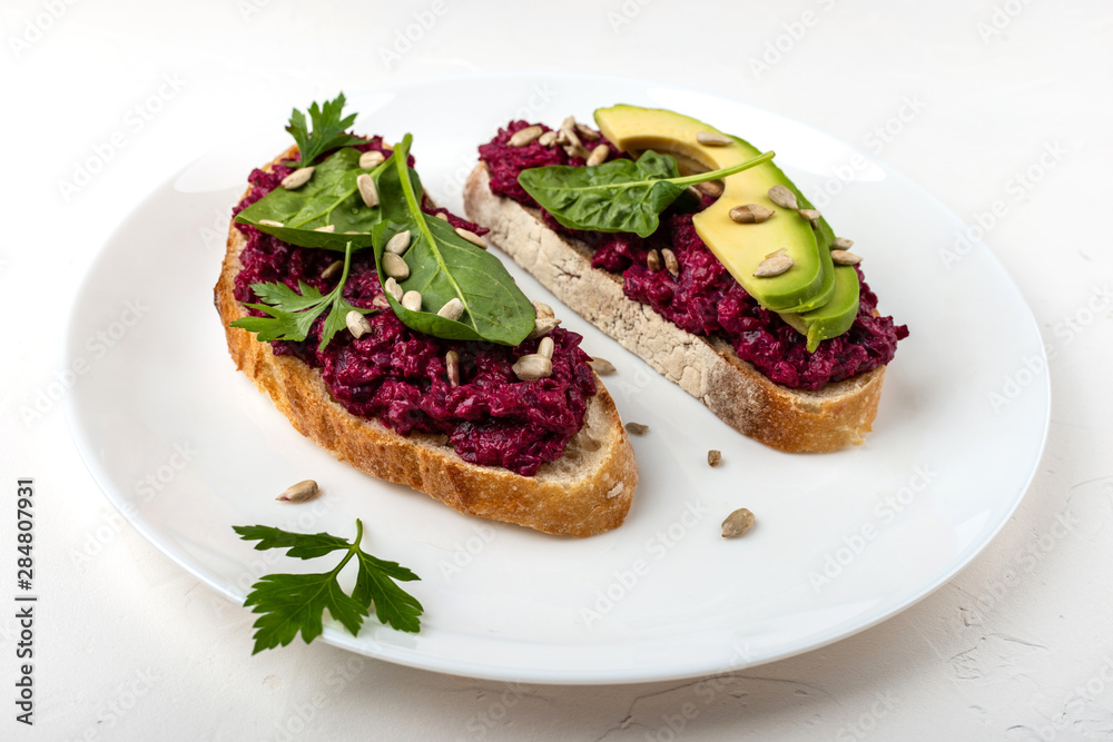 Bruschettas with beetroot hummus, avocado, spinach and seeds on a white plate on a white background. Bruschettas with beetroot hummus, avocado, spinach and seeds on a white plate on a white background.