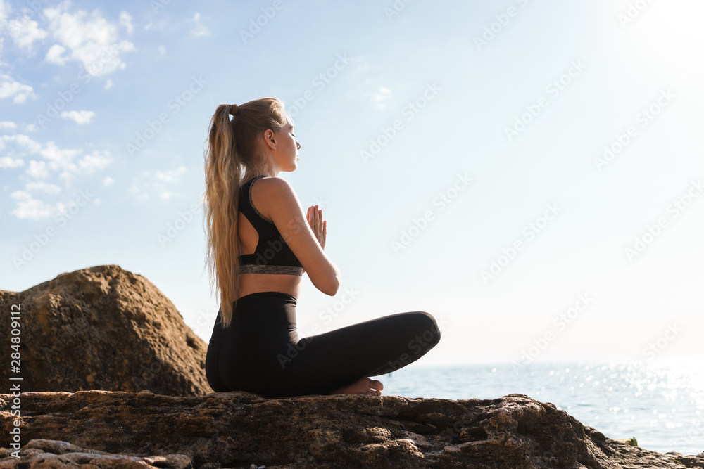 Image of calm concentrated woman meditating while sitting on rock at seaside in morning