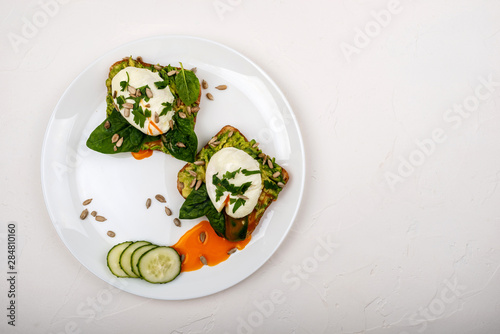Poached eggs with avocado, spinach leaves and seeds on toasts bread on a white plate on the white background. Healthy breakfast or snack. Cope space.