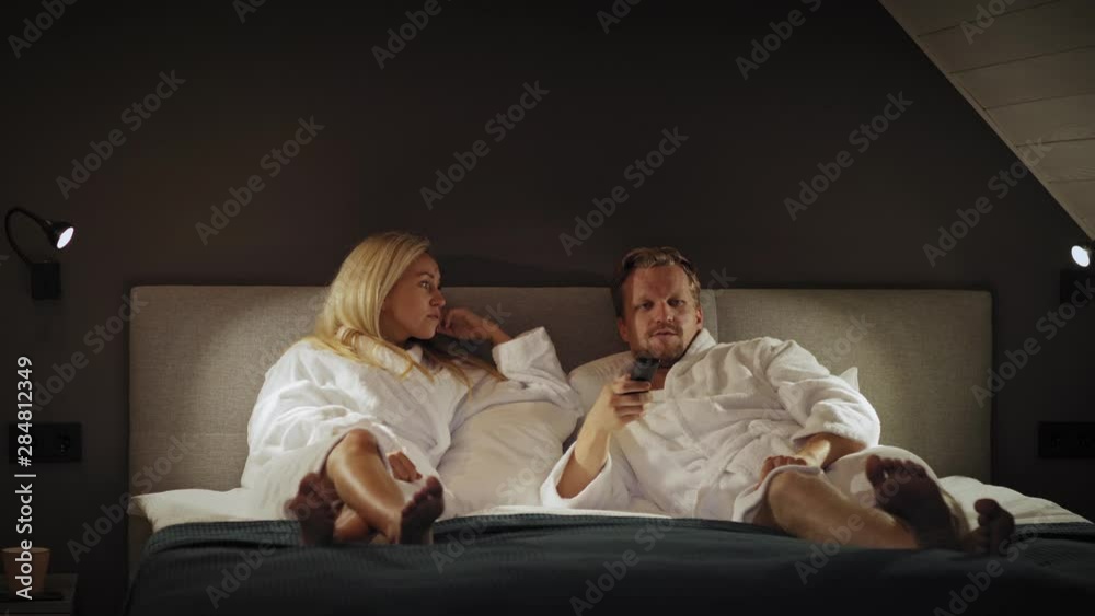 Emotional middle aged couple in white bathrobes lying on bed in hotel room or at home and fighting for remote control to switch channels at night Emotional middle aged couple in white bathrobes lying on bed in hotel room or at home and fighting for remote control to switch channels at night