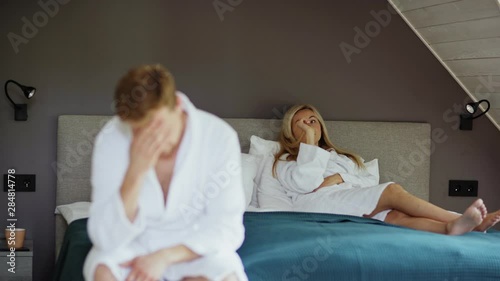 Upset woman in white bathrobe lying on bed and touching her hair anxiously. Defocused depressed husband or lover rubbing forehead in foreground