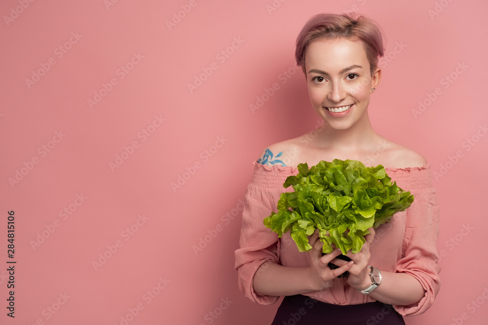 A girl with short pink hair smiling looking at the camera, holding a salad near her chest, on a pink background. A girl with short pink hair smiling looking at the camera, holding a salad near her chest, on a pink background.