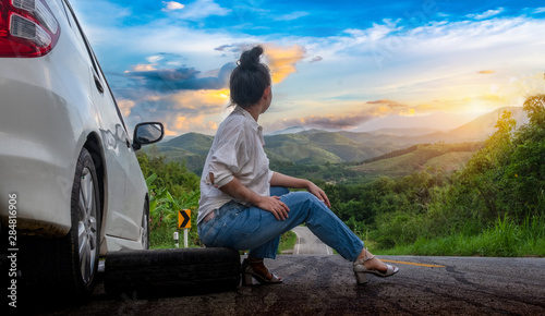Young beautiful lady sit near car for calling for help on the public road in forest area