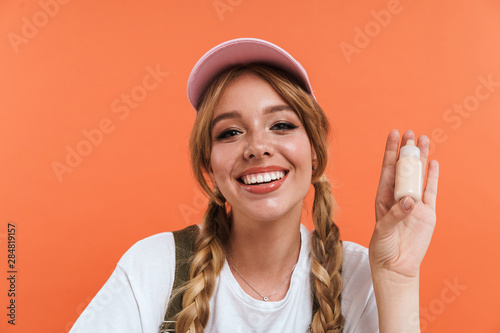 Image of joyful blogger girl wearing hat holding cosmetic bottle and demonstrating new product