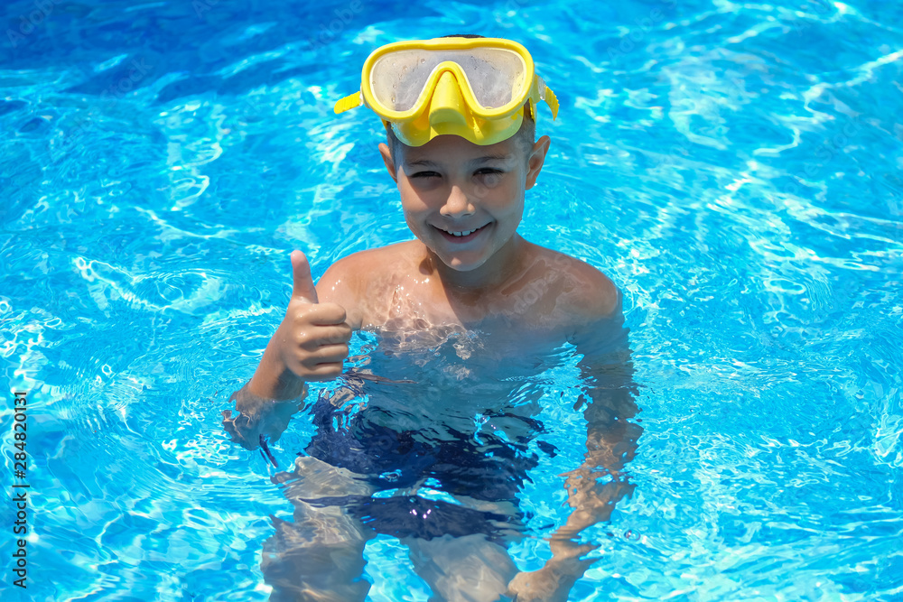 A child in a mask for swimming. A young boy in the pool shows thumbs up. Kid are engaged in diving.