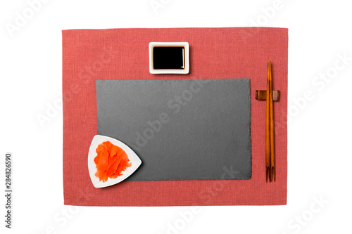Empty rectangular black slate plate with chopsticks for sushi, ginger and soy sauce on red napkin background. Top view with copy space for you design