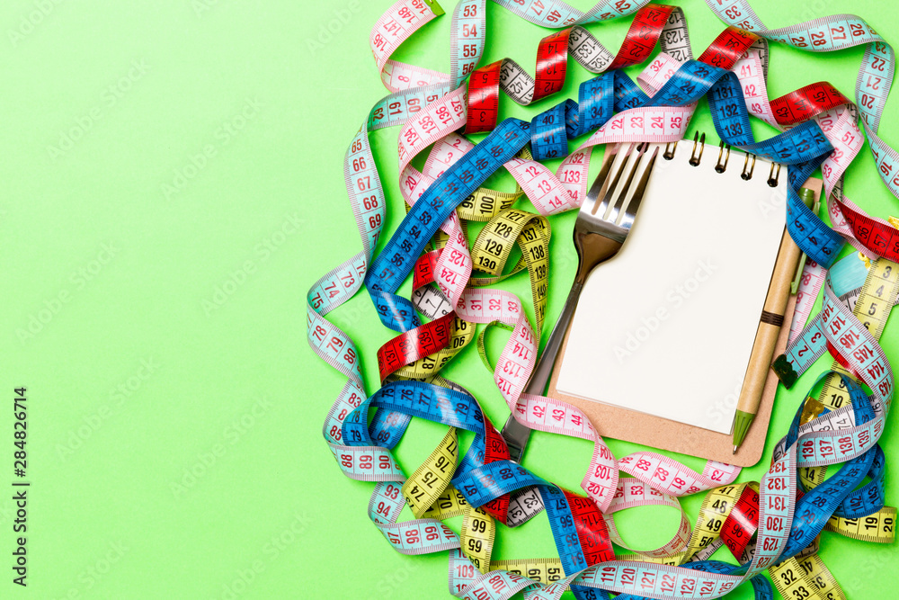 Fototapeta premium Composition of colorful measuring tapes, fork, pen and notepad on green background. Top view of diet control and empty space for your design