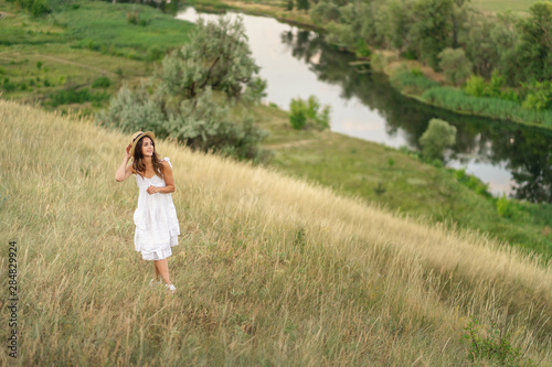 Smiling woman in white dress and straw hat is walking near the river. Summer nature. Meadow