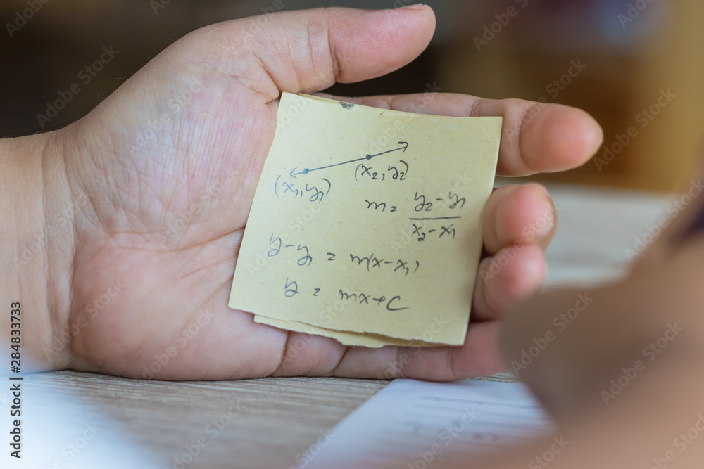 Student using formula math sheet to cheat on test. Hand of university student holding pencil doing math question in examination and cheating with answer piece of paper in hand. Education concept. Student using formula math sheet to cheat on test. Hand of university student holding pencil doing math question in examination and cheating with answer piece of paper in hand. Education concept.