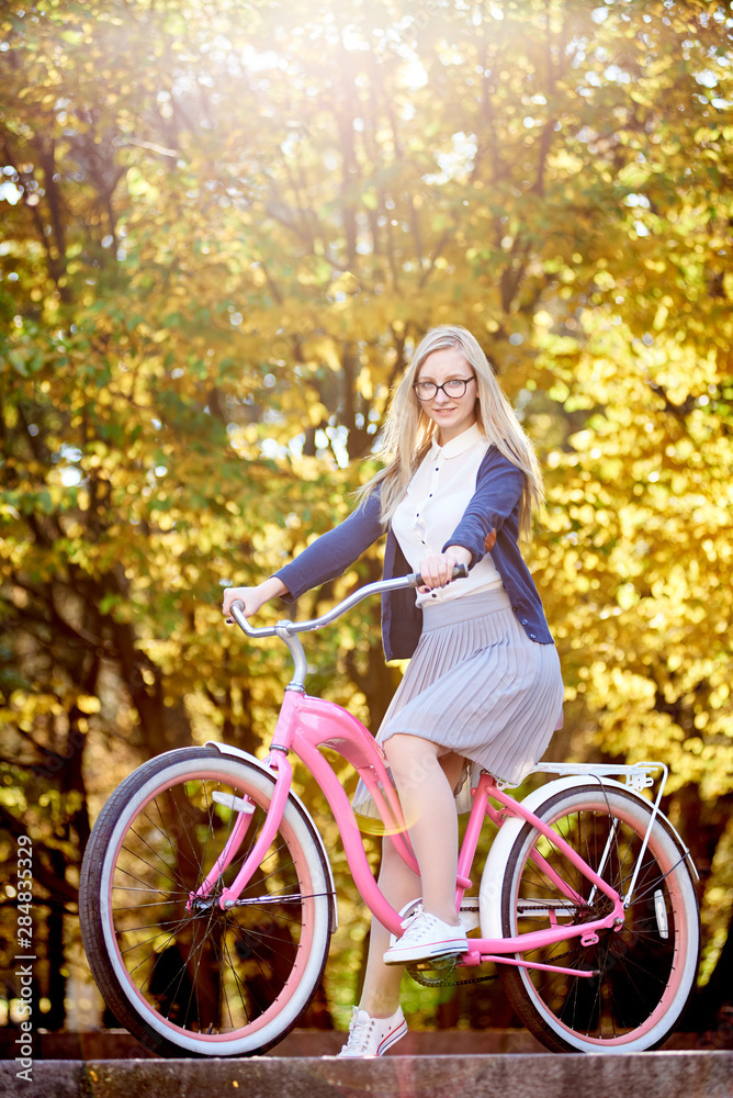 Active blond long-haired attractive traveler girl in glasses, skirt and blouse posing on modern pink lady bicycle on lit by autumn sun park stairs on bright colorful golden bokeh trees background. Active blond long-haired attractive traveler girl in glasses, skirt and blouse posing on modern pink lady bicycle on lit by autumn sun park stairs on bright colorful golden bokeh trees background.