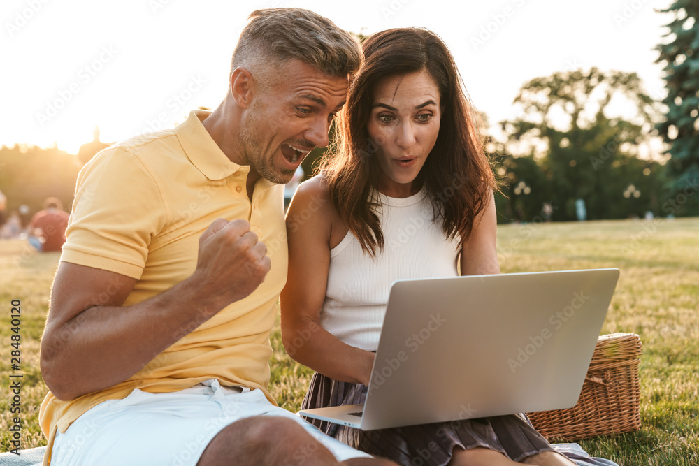 Portrait of surprised middle-aged couple man and woman celebrating success while using laptop computer in summer park
