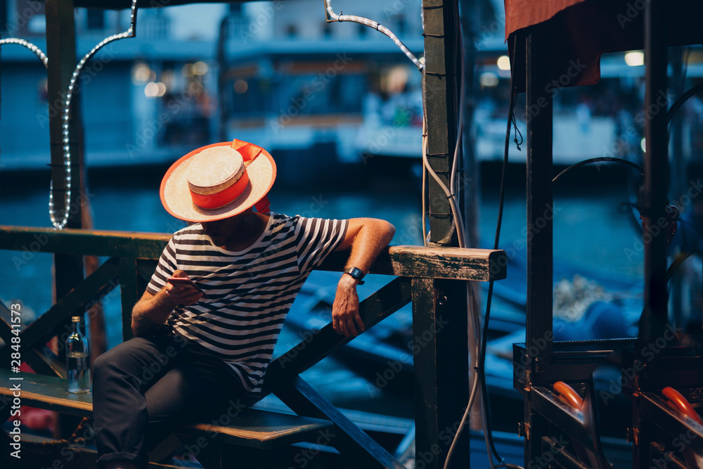 Φωτογραφία Gondolier in  hat and striped clothes sits in  evening on canal and waits for cl