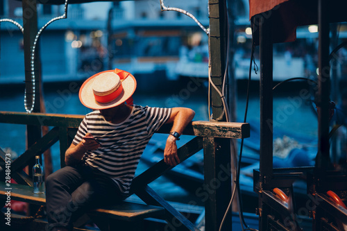 Photo Gondolier in  hat and striped clothes sits in  evening on canal and waits for cl