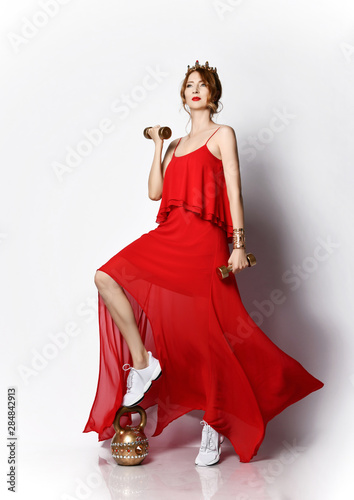 Fitness queen sporty woman in long red dress and sneakers stands with her foot on weight and workouts on white