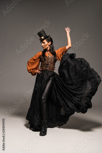 full length view of attractive steampunk woman in top hat with goggles standing with hand on hip on grey