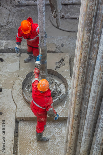 Two working drillers in a red uniform, in a helmet and goggles, install drill pipes after lifting them from an oil well after drilling. The concept of a working person. photo