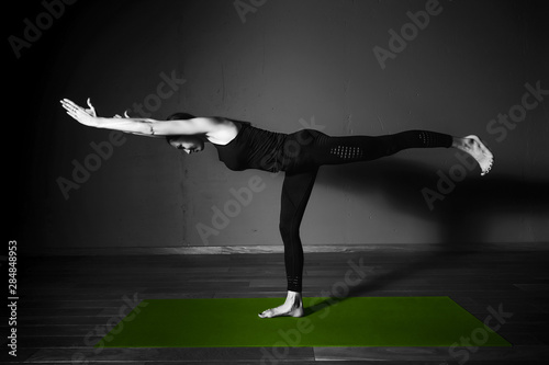 Black white photo of Young beautiful woman brunette in black clothes practice yoga virabhadrasana 3 in dark studio green mat Sport meditation monochrome photo