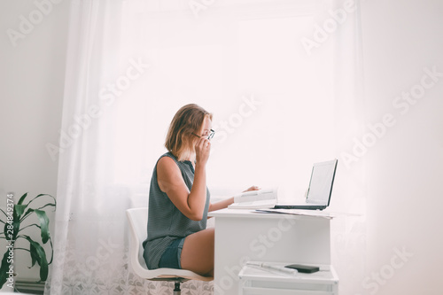 Young woman with glasses sitting at the table and leafing through a book. Nearby are a laptop and office supplies. Interior is white. Copy space © _KUBE_