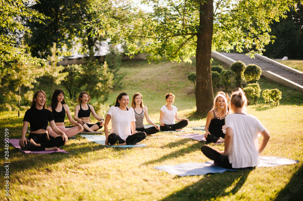 Group of smiling women prepare to meditation in park on summer sunny morning under guidance of trainer. Group of girl are sitting in lotus pose on yoga mats are relaxing talking before yoga training - obrazy, fototapety, plakaty 