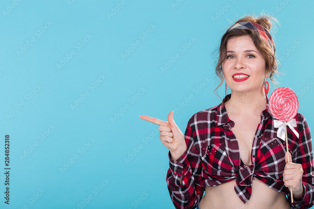 Smiling beautiful young girl in a plaid shirt with a lollipop in her hands posing on a blue background and shows a finger to the right. Concept information and subscription link. Copy space Smiling beautiful young girl in a plaid shirt with a lollipop in her hands posing on a blue background and shows a finger to the right. Concept information and subscription link. Copy space