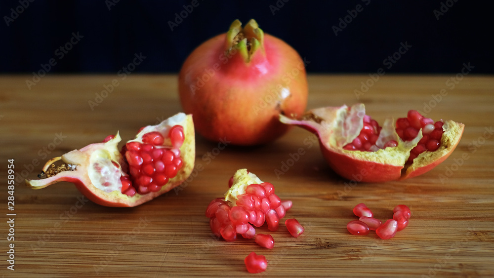 One whole and one open pomegranate or the fruit of Punica granatum revealing clusters of red, juicy seeds, white fibrous arils and thick, waxy peel resting on a wooden board with a dark background One whole and one open pomegranate or the fruit of Punica granatum revealing clusters of red, juicy seeds, white fibrous arils and thick, waxy peel resting on a wooden board with a dark background