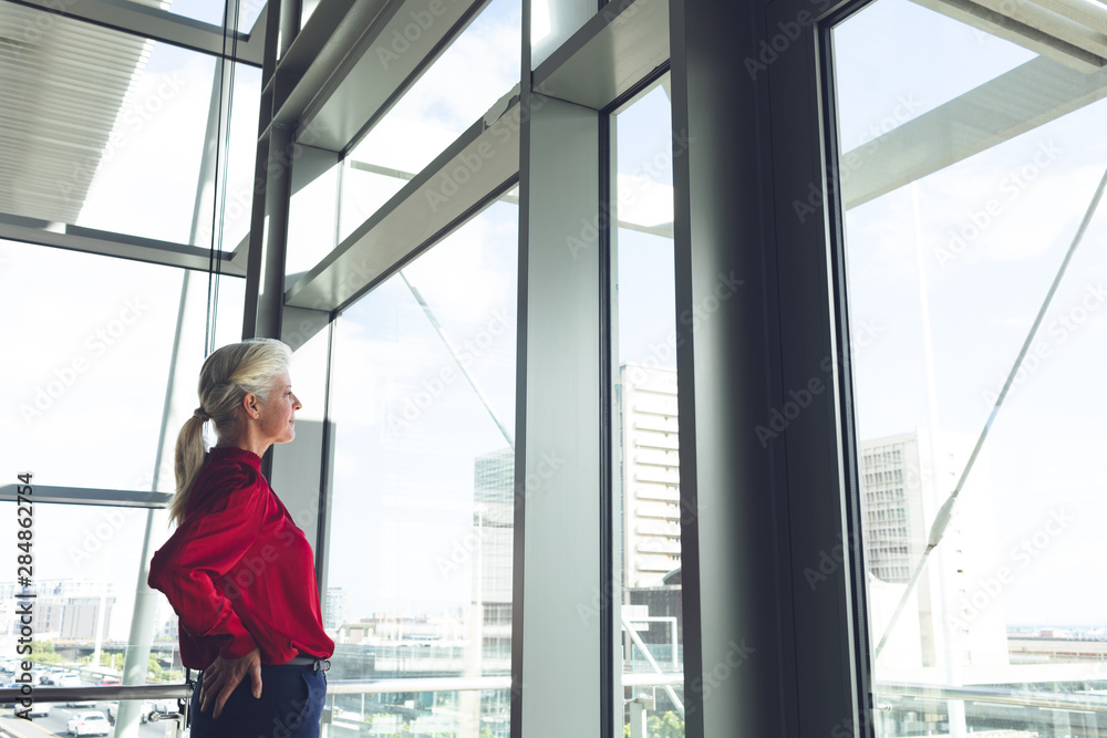Senior businesswoman with hands on hip looking through window in office building Senior businesswoman with hands on hip looking through window in office building