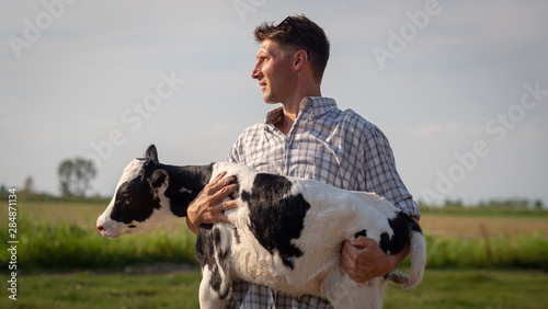 Fotografia Authentic shot of young man farmer is holding on his arms an ecologically grown newborn calf used for biological milk products industry on a green lawn of a countryside farm