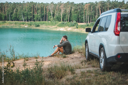 man sitting near white suv car at the edge looking at lake with blue water