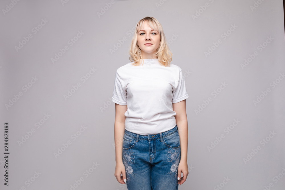 View from front of beautiful slim woman wearing white shirt and jeans standing steady on frey isolated background. Young blonde looking at camera, smiling and posing in studio. View from front of beautiful slim woman wearing white shirt and jeans standing steady on frey isolated background. Young blonde looking at camera, smiling and posing in studio.