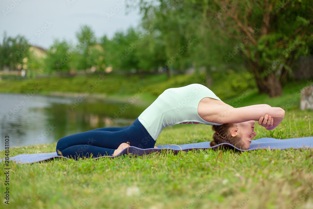 Thin brunette girl plays sports and performs beautiful and sophisticated yoga poses in a summer park. Green lush forest and the river on the background. Woman doing exercises on a yoga mat