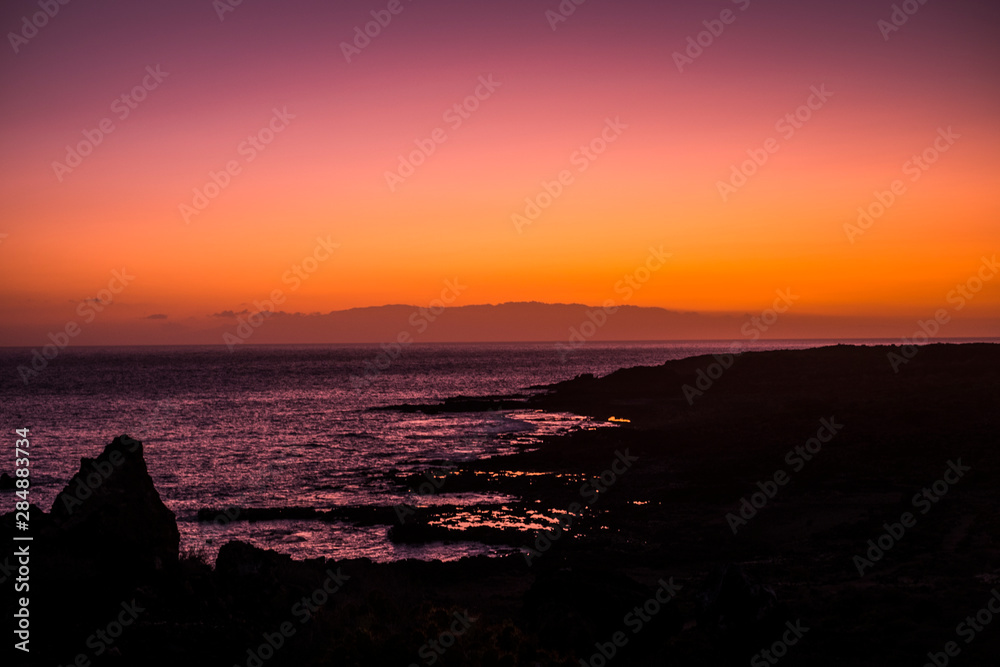 Romantic scenic sunset on the coast with black. rock beach and quiet wave ocean - sky and clouds in background - red and orange colours in landscape nature outdoor