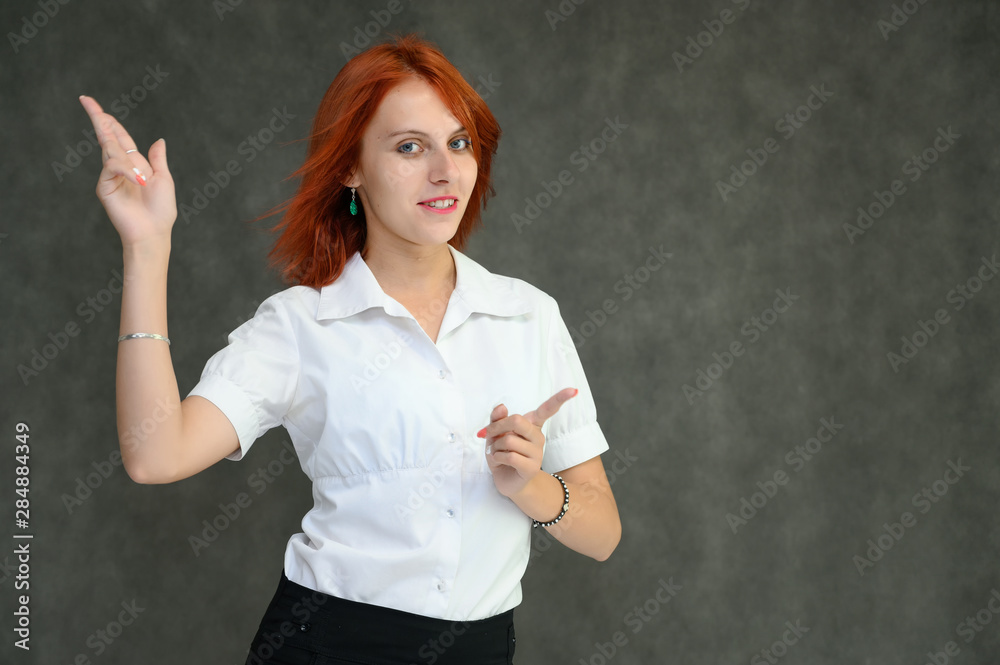 Photo Portrait of a cute girl woman with bright red hair manager in a white shirt on a gray background in the studio. He talks, shows his hands in front of the camera with emotions. Photo Portrait of a cute girl woman with bright red hair manager in a white shirt on a gray background in the studio. He talks, shows his hands in front of the camera with emotions.