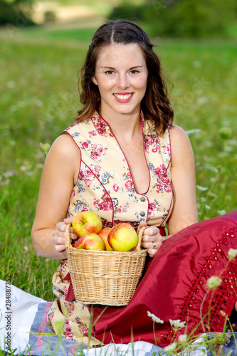 woman in drindl sitting in a meadow and holding a basket with apples photo