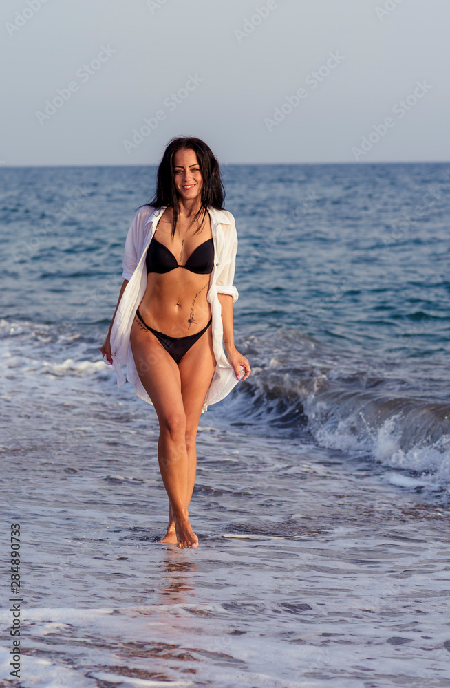 sexy and brunette woman with black bikini and white shirt walking along the seashore. Summer time concept.
