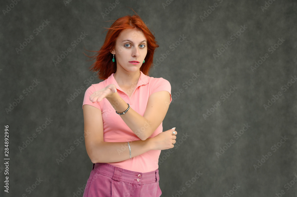 Photo Portrait of a cute woman girl with bright red hair in a peach t-shirt and pink skirt on a gray background in the studio. He talks, shows his hands in front of the camera with emotions. Photo Portrait of a cute woman girl with bright red hair in a peach t-shirt and pink skirt on a gray background in the studio. He talks, shows his hands in front of the camera with emotions.