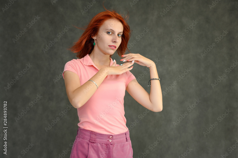 Fototapeta premium Photo Portrait of a cute woman girl with bright red hair in a peach t-shirt and pink skirt on a gray background in the studio. He talks, shows his hands in front of the camera with emotions.