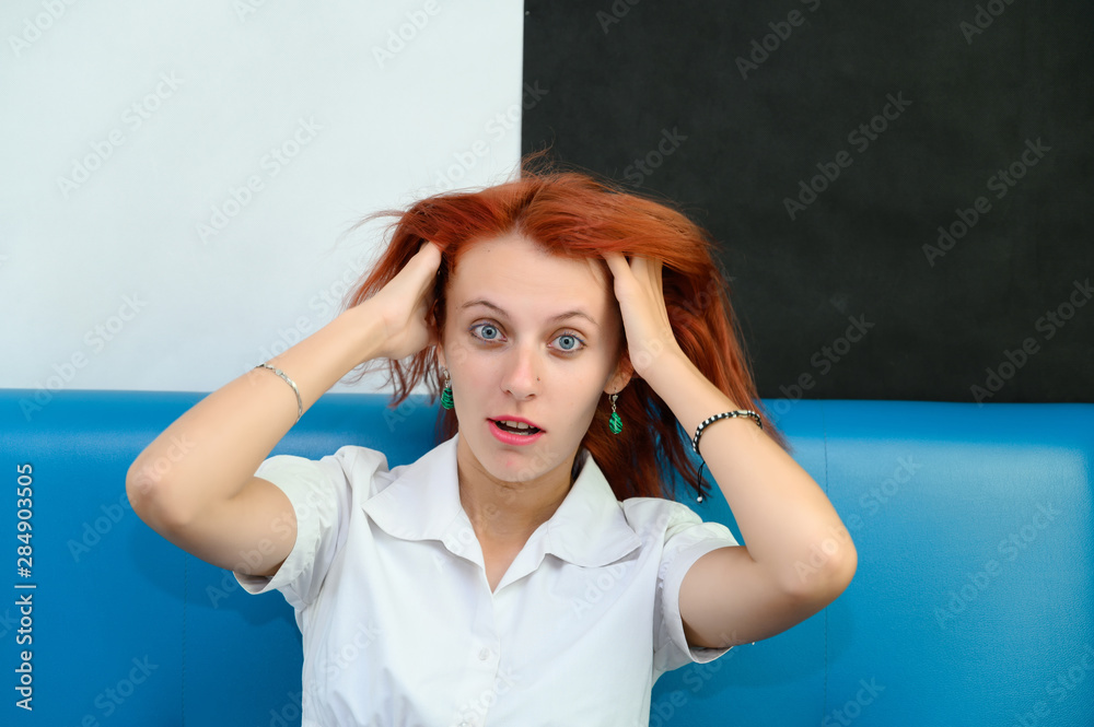Photo Portrait of a cute woman girl with bright red hair in a white T-shirt on a black and white background in studio. Sits on a blue sofa, Talks in front of the camera with emotions.