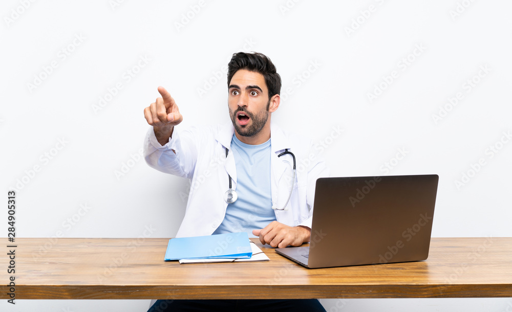 Young doctor man with his laptop over isolated wall pointing away Young doctor man with his laptop over isolated wall pointing away