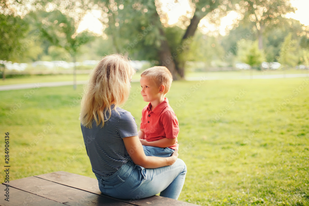 Fototapeta premium Young blonde Caucasian mother and boy toddler son sitting together face to face on wooden bench outdoor on summer day and talking to each other. Happy family childhood lifestyle.