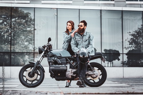 Portrait of attractive couple in denim jackets with motorbike near big glass building at city centre. photo