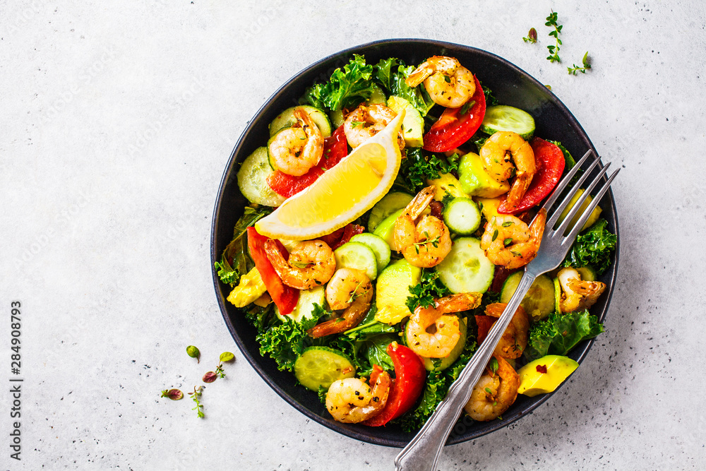 Shrimp, avocado and vegetable salad in a black dish on gray background, top view.