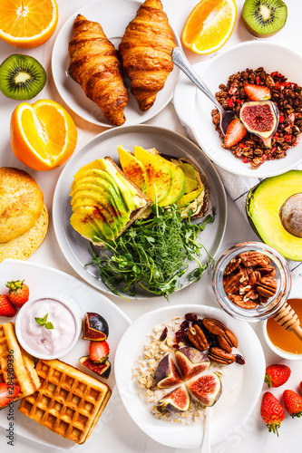 Breakfast table with avocado toast, oatmeal, waffles, croissants on a white background.