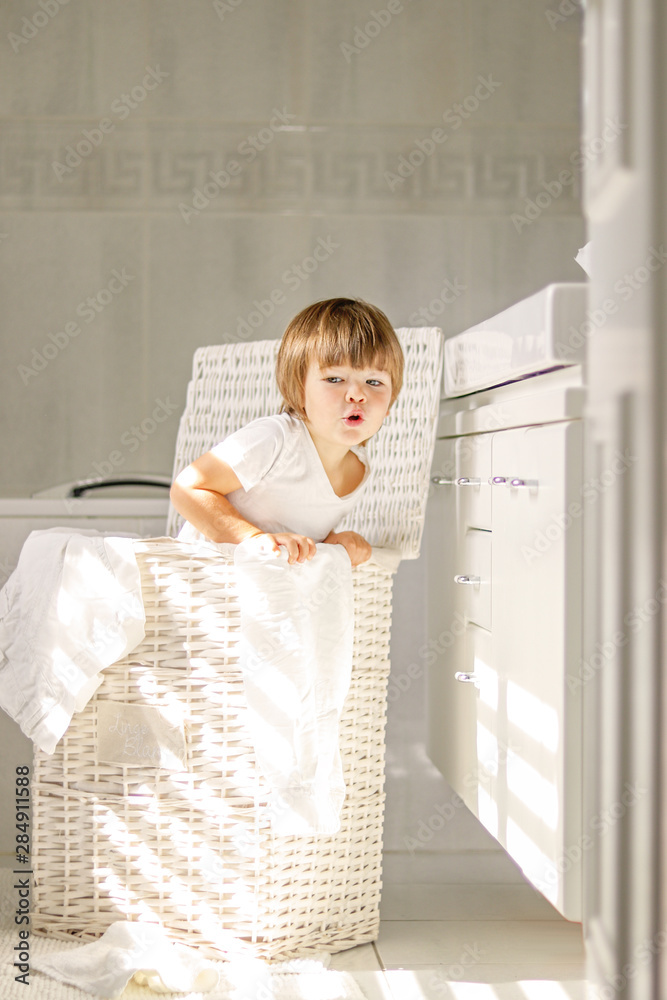 Cute little playful boy with funny face expression standing inside white lwicker laundry basket with clothes hanging out in white bathroom with natural light. Happy child helping mom. Cute little playful boy with funny face expression standing inside white lwicker laundry basket with clothes hanging out in white bathroom with natural light. Happy child helping mom.