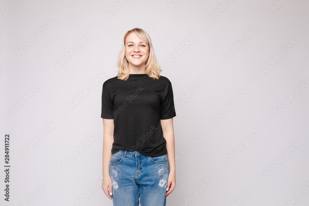 View from front of beautiful slim woman wearing black shirt and jeans standing steady on frey isolated background. Young blonde looking at camera, smiling and posing in studio. View from front of beautiful slim woman wearing black shirt and jeans standing steady on frey isolated background. Young blonde looking at camera, smiling and posing in studio.