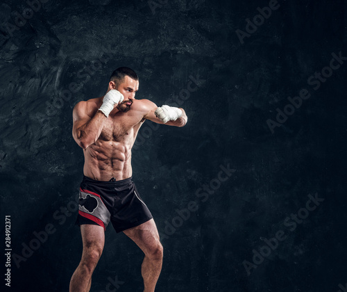 Strong muscular fighter is showing his punch while posing for photographer at dark photo studio. © Fxquadro